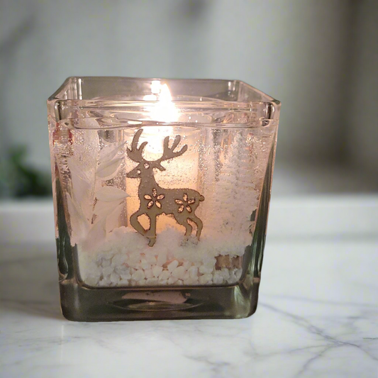 Photophore en verre carré avec un cerf, une bougie allumée à l’intérieur et des décorations blanches ressemblant à de la neige au fond, placé sur un comptoir en marbre, créant une ambiance festive et chaleureuse.